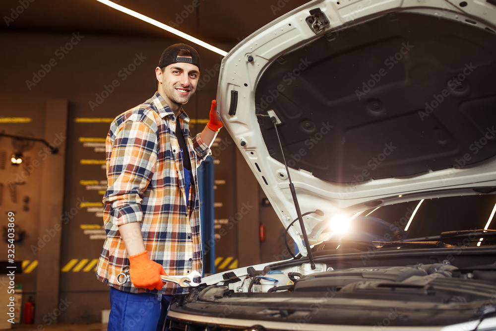 auto mechanic holding wrenches with open car hood looking at the camera auto mechanic holding wrenches with open car hood looking at the camera