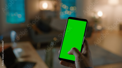 Close Up Shot of a Smartphone with Green Screen Great for Mock-up. Person is Holding Phone and Giving a Voice Command to Switch on the Light on the Backgroung in Living Room.