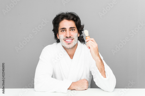 Young caucasian man shaving his beard isolated on a grey background