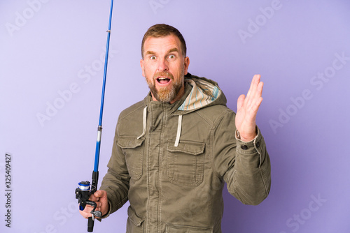 Senior fisherman isolated on purple background receiving a pleasant surprise, excited and raising hands.