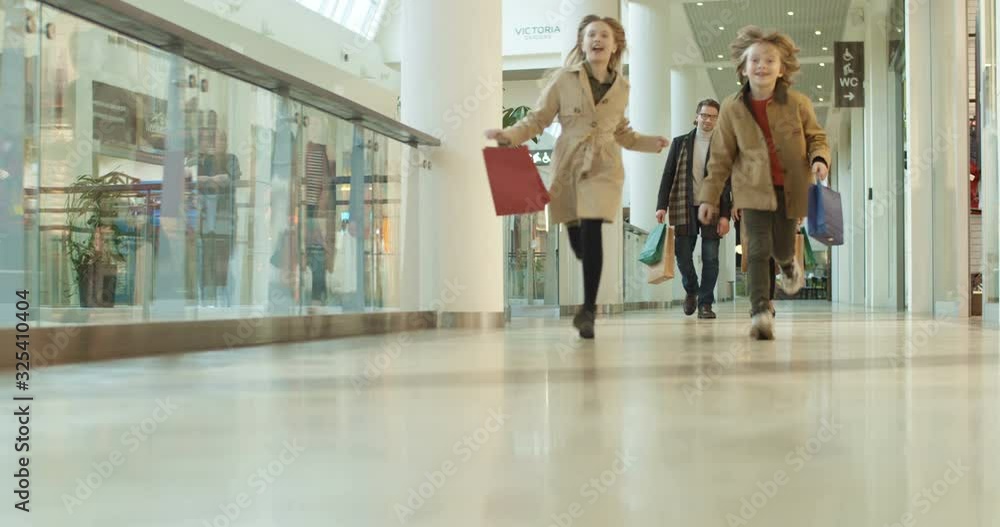 Happy Caucasian kids sister and brother running shopping mall passage with packets and parents following them by walking. Cheerful children having fun in store with mother and father. Happy Caucasian kids sister and brother running shopping mall passage with packets and parents following them by walking. Cheerful children having fun in store with mother and father.