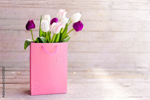 Spring flowers. Blooming tulips in pink shopping bag at wooden background. White, lilac and purple bouquet. Still life in morning sun light. Presents for holidays. Copy space.