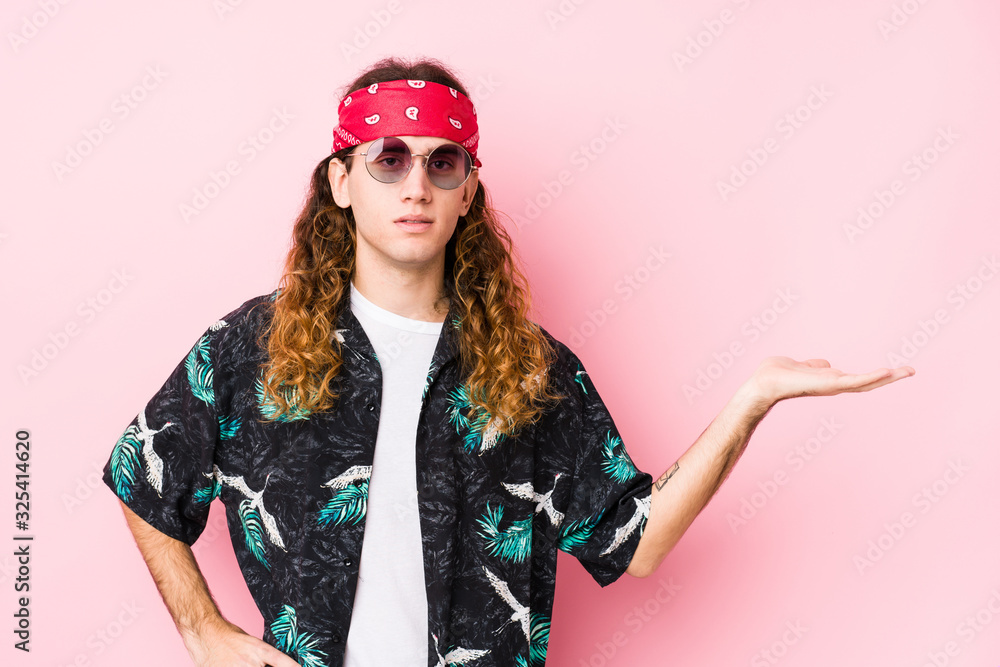 Young hippie caucasian man isolated showing a copy space on a palm and holding another hand on waist.