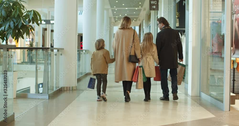 Rear of Caucasian family with kids spending weekend in shopping mall. Smiled parents walking hand in hand with son daughter with bags. Back view on little sister and brother with mother and father. Rear of Caucasian family with kids spending weekend in shopping mall. Smiled parents walking hand in hand with son daughter with bags. Back view on little sister and brother with mother and father.