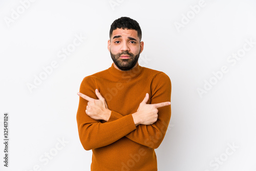 Young latin man against a white background isolated points sideways, is trying to choose between two options.