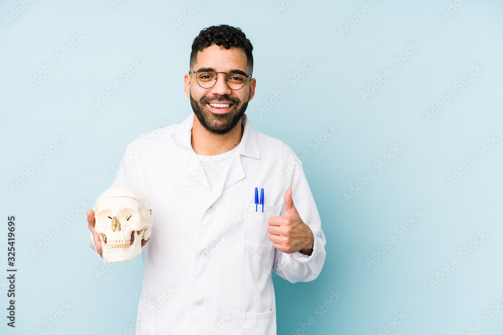 Young doctor latin man holding a skull isolated smiling and raising thumb up Young doctor latin man holding a skull isolated smiling and raising thumb up