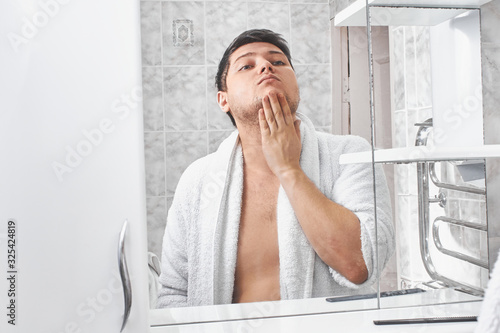 a man in a white bathrobe looks in the mirror in the bathroom.