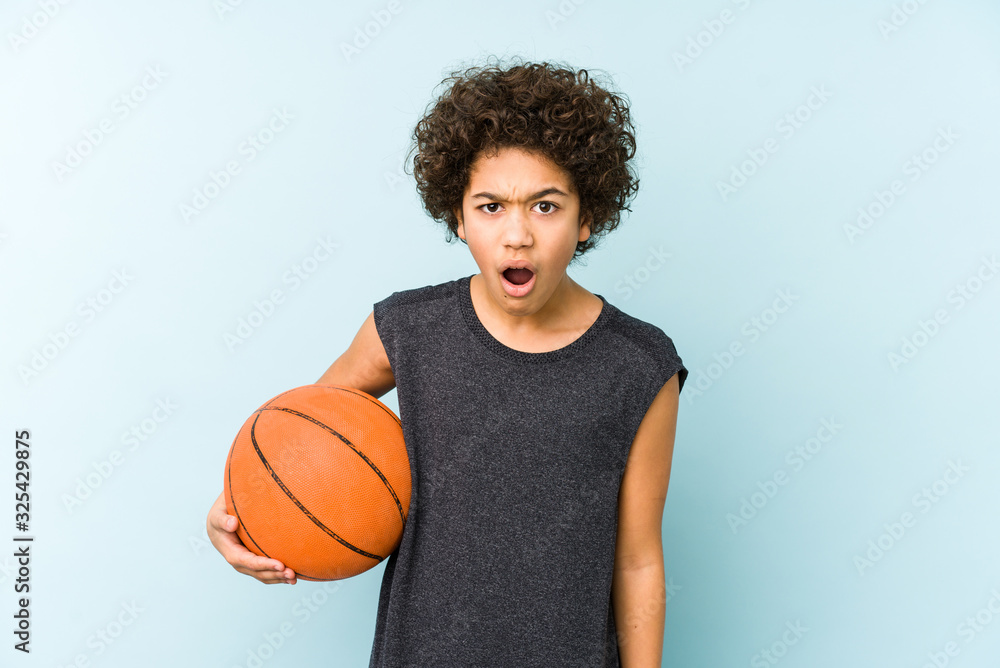 Kid boy playing basketball isolated on blue background screaming very angry and aggressive.