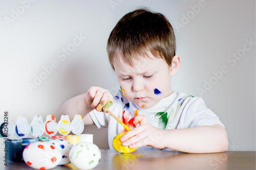 Beautiful little child sits at a table and draws Easter eggs with bright paint. Boy on a light background in a white T-shirt, face, clothes and hands painted with colorful paint