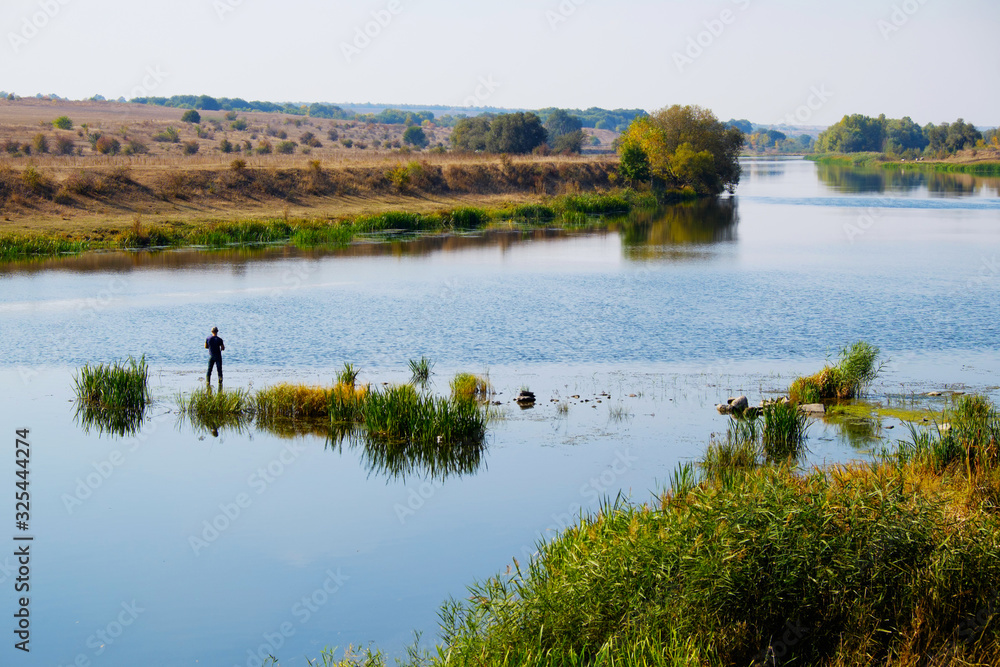 Man traveler silhouette near river with green forest hills. Travel outdoor lifestyle. Fisherman standing in grass, fishing and enjoy autumn nature. Rural landscape with blue lake and man catch fish.