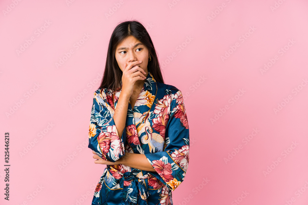Young asian woman wearing a kimono pajamas thoughtful looking to a copy space covering mouth with hand. Young asian woman wearing a kimono pajamas thoughtful looking to a copy space covering mouth with hand.