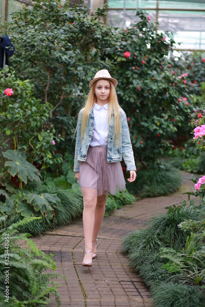 Young girl with long white hair in a  hat and a denim jacket walks in the garden with blooming azalea