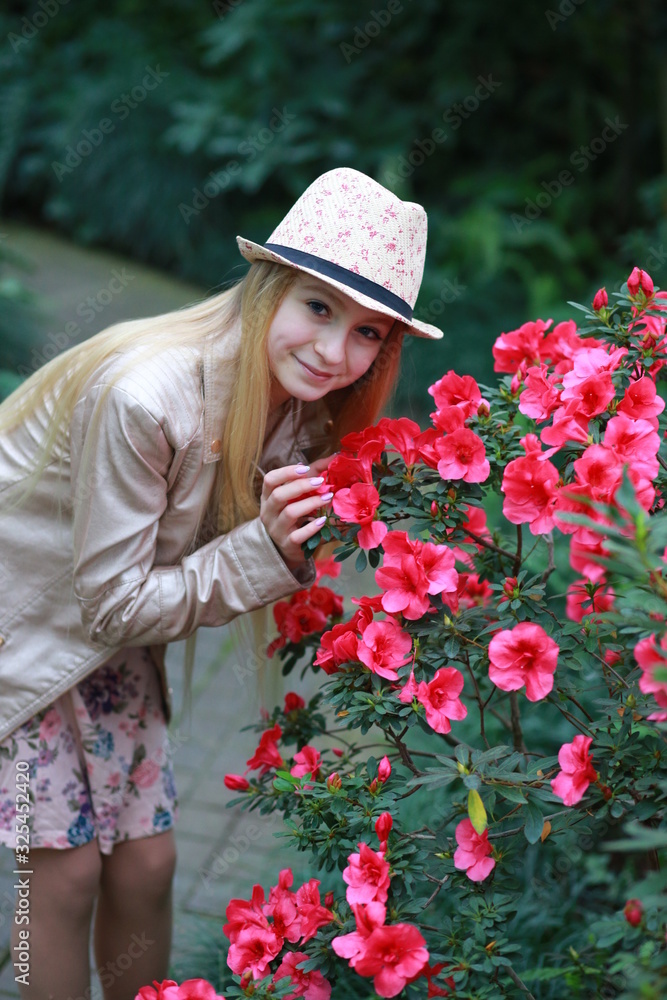 Young girl with long blond hair in a powdery jacket and a hat enjoys in the garden with blooming azalea