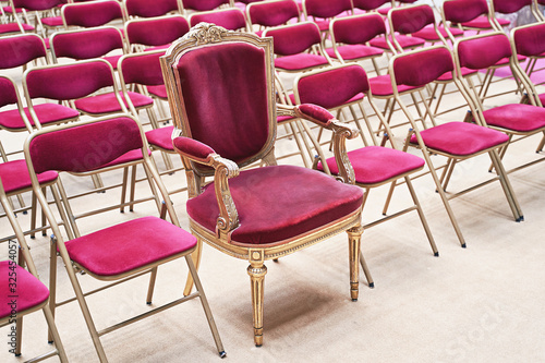 Unique throne or ceremonial armchair with velvet seat and golden details among many simple identical similar chairs. Uniqueness or exclusivity concept.