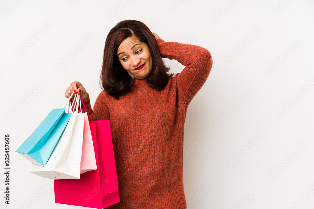 Middle age latin woman holding a shopping bags isolated touching back of head, thinking and making a choice. Middle age latin woman holding a shopping bags isolated touching back of head, thinking and making a choice.