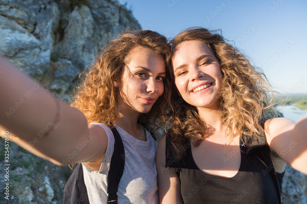 Fototapeta premium A world without borders. Stunning journey of two girls in the mountains. two attractive young girls travel together, do selfie on the phone against the sky and landscape in the mountains in the sun