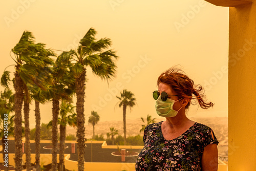 A woman stands on a balcony in the hotel and has a face mask and sunglasses to protect her from the Calima sandstorm, which brings a lot of fine dust. Concept: health and travel photo