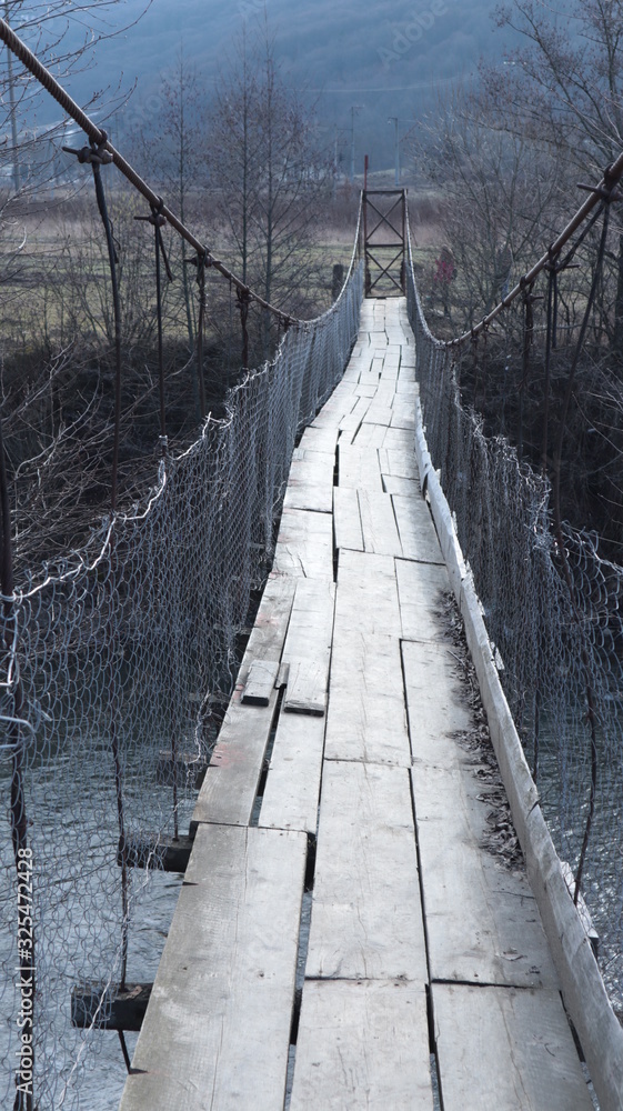 Fototapeta premium old pedestrian suspension bridge over a mountain river of metal bars and wooden floor