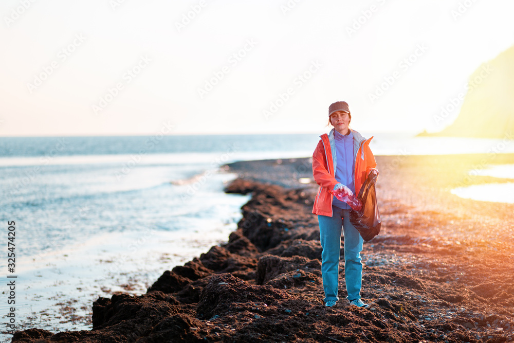 A young Caucasian woman in a jacket collects garbage and puts it in a plastic bag. In the background, the beach after the storm and the sea. Copy space