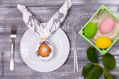 Composition with an Easter egg wrapped in a napkin in the form of rabbit ears on a white plate, cutlery, colorful Easter eggs in a green wooden box on a wooden gray table. Flat lay