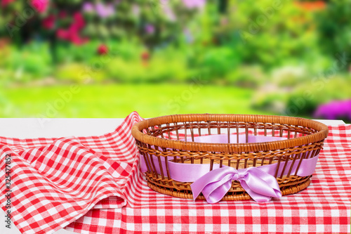 Table top on empty basket background. Empty picnic basket with pink ribbon on red checkered tablecloth with natural blurred summer garden background. For your food and product display montage.