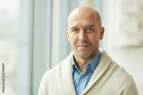 Head and shoulders portrait of balding mature man wearing cardigan looking at camera while standing by window in white office, copy space