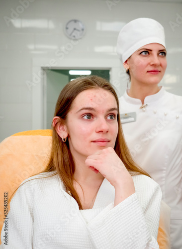 A young girl with problem skin is seated on a couch before cosmetic procedures at the cosmetology clinic. Beautician in the background out of focus