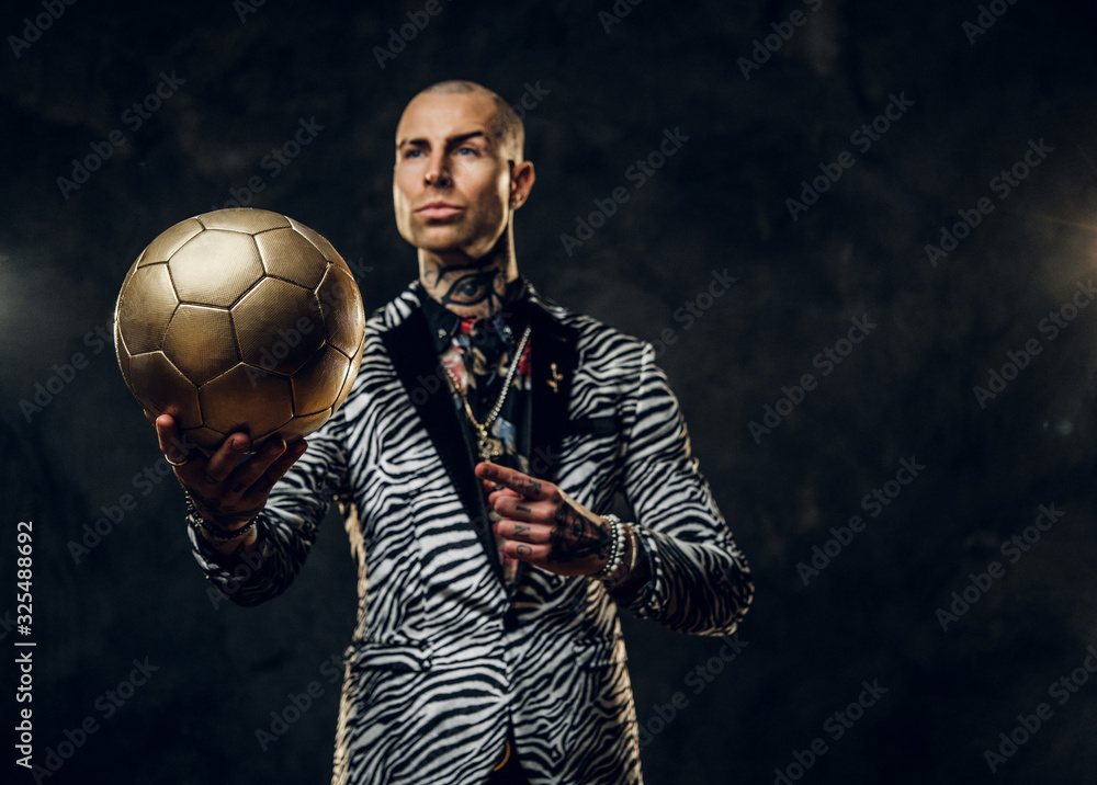 Plakat Elegant, vivid, tattooed, bald male model posing in a studio for the photoshoot wearing fashionable custom made zebra striped style tuxedo and rose patterned shirt, looking on a golden soccer ball