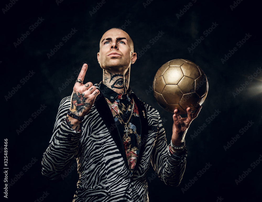 Plakat Elegant, vivid, tattooed, bald male model posing in a studio for the photoshoot wearing fashionable custom made zebra striped style tuxedo and rose patterned shirt, looking on a golden soccer ball
