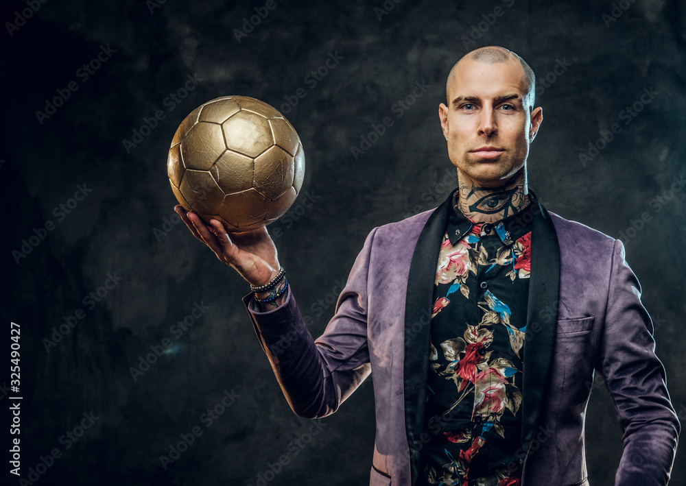 Plakat Handsome, sporty, tattooed, bald male model posing in a studio for the photoshoot wearing fashionable purple tuxedo and rose patterned shirt, looking calm on camera while holding a golden soccer ball
