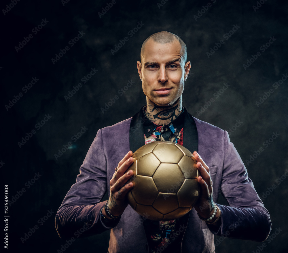 Plakat Handsome, sporty, tattooed, bald male model posing in a studio for the photoshoot wearing fashionable purple tuxedo and rose patterned shirt, looking expressive on camera while holding a golden soccer