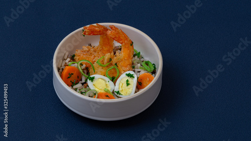 Rice bowl with vegetables, shrimps and eggs. There is leek and carrots in it. White bowl, blue background.