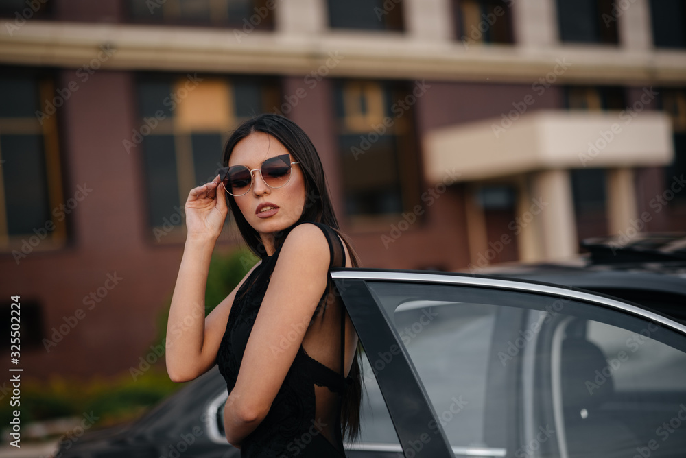 Stylish young girl stands near the car in a black dress. Business fashion and style