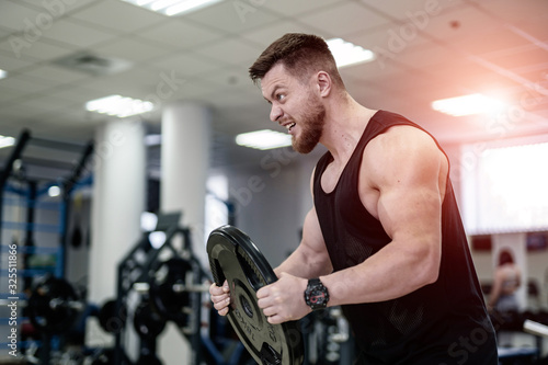 Portrait of handsome muscular powerlifter with weight disk in his hands in the sport gym. Bodybuilder with heavy disk trains his biceps with the open mouth. Close-up