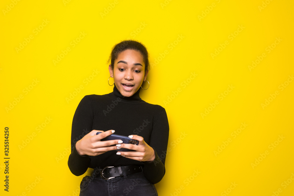 Portrait of a cheerful young latin woman playing games on mobile phone isolated over yellow background Portrait of a cheerful young latin woman playing games on mobile phone isolated over yellow background