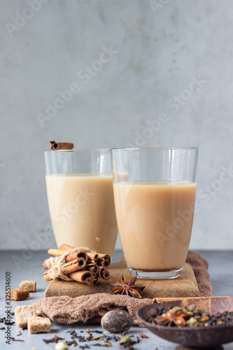 Two drinking glasses with traditional Indian drink - masala chai tea (milk tea) with spices on grey stone background with spices for making tea.