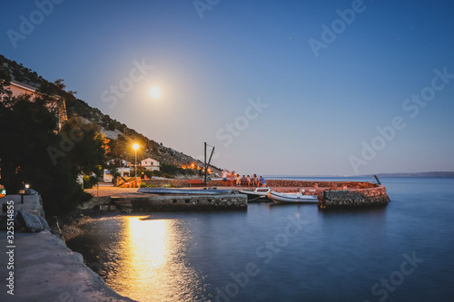 Typical croatian bay on the coast of Velebit or Kvarner during a calm summer night. Moon is visible above the people sitting on the marina wall.
