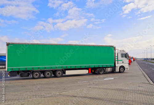 A row of trucks in a parking lot. In the foreground a truck with a semi-trailer. Green tarpaulin.