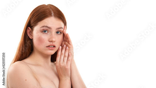 Fresh and clean. Portrait of a young sensual red-haired girl with freckles touching her face and looking at camera while standing against white background
