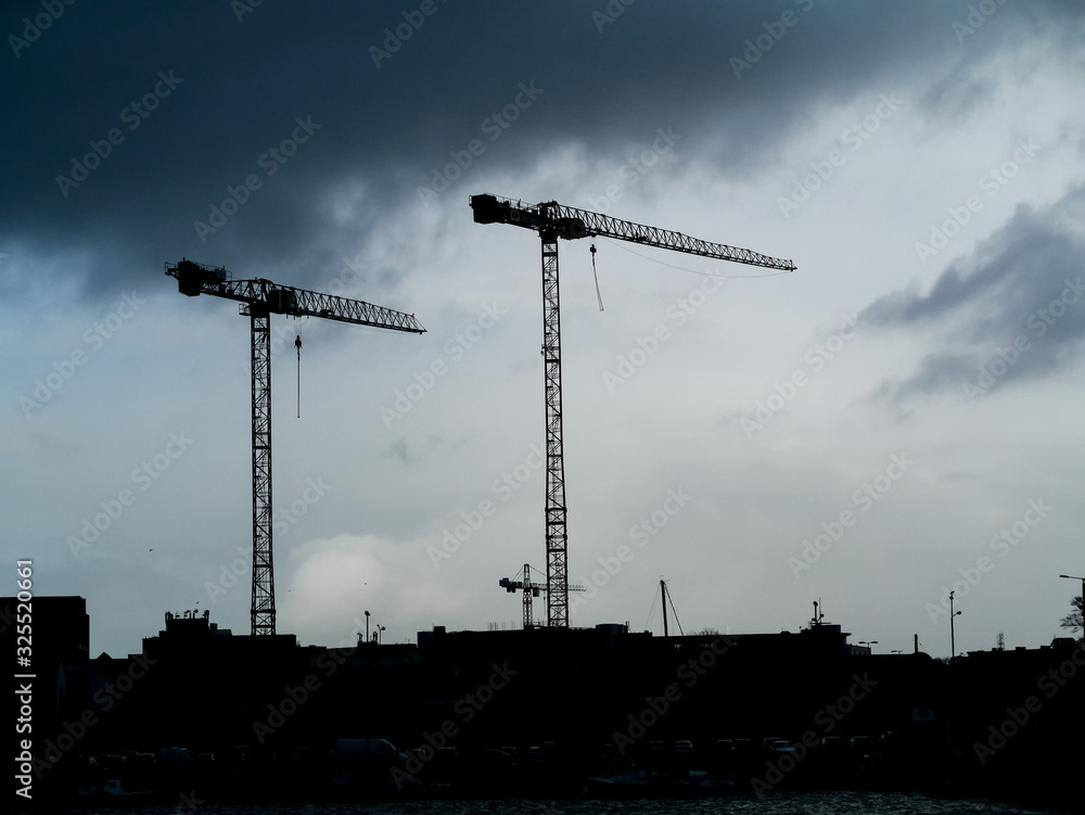 Two heavy tall cranes on a construction site against cloudy sky. Two heavy tall cranes on a construction site against cloudy sky.