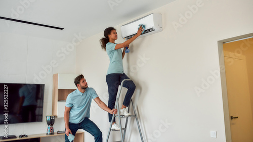 Service at work. Young afro american woman in uniform cleaning the air conditioner while standing on the ladder in the living room. Two professional cleaners working together
