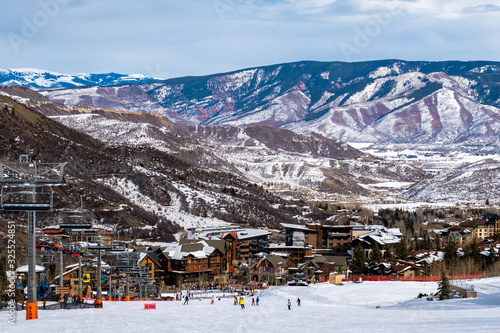 Panoramic view of Snowmass Village, with skiers skiing at the Aspen Snowmass ski resort in the foreground and the Rocky Mountains of Colorado in the background, on a partly cloudy winter day.   © David A Litman