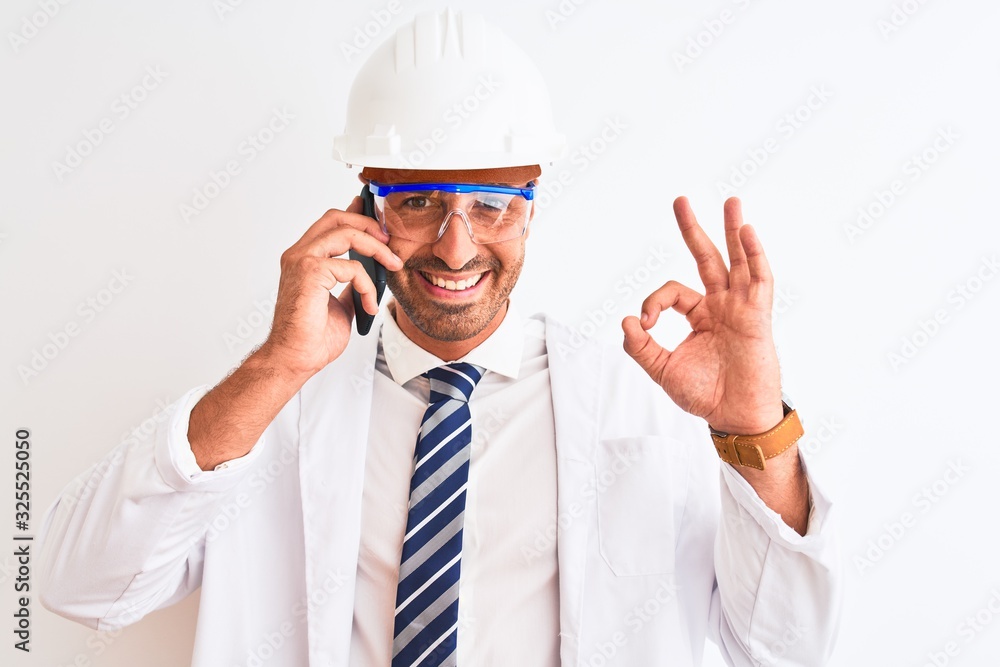 Young scientist man wearing safety helmet and calling using smartphone over isolated background doing ok sign with fingers, excellent symbol Young scientist man wearing safety helmet and calling using smartphone over isolated background doing ok sign with fingers, excellent symbol
