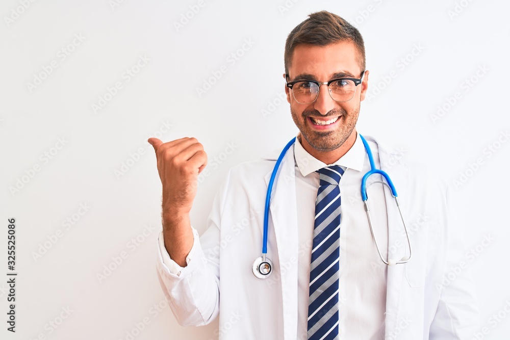 Young handsome doctor man wearing stethoscope over isolated background smiling with happy face looking and pointing to the side with thumb up. Young handsome doctor man wearing stethoscope over isolated background smiling with happy face looking and pointing to the side with thumb up.