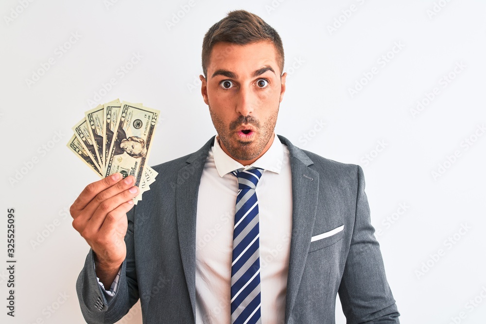 Young handsome business man holding bunch of dollars banknotes over isolated background scared in shock with a surprise face, afraid and excited with fear expression Young handsome business man holding bunch of dollars banknotes over isolated background scared in shock with a surprise face, afraid and excited with fear expression