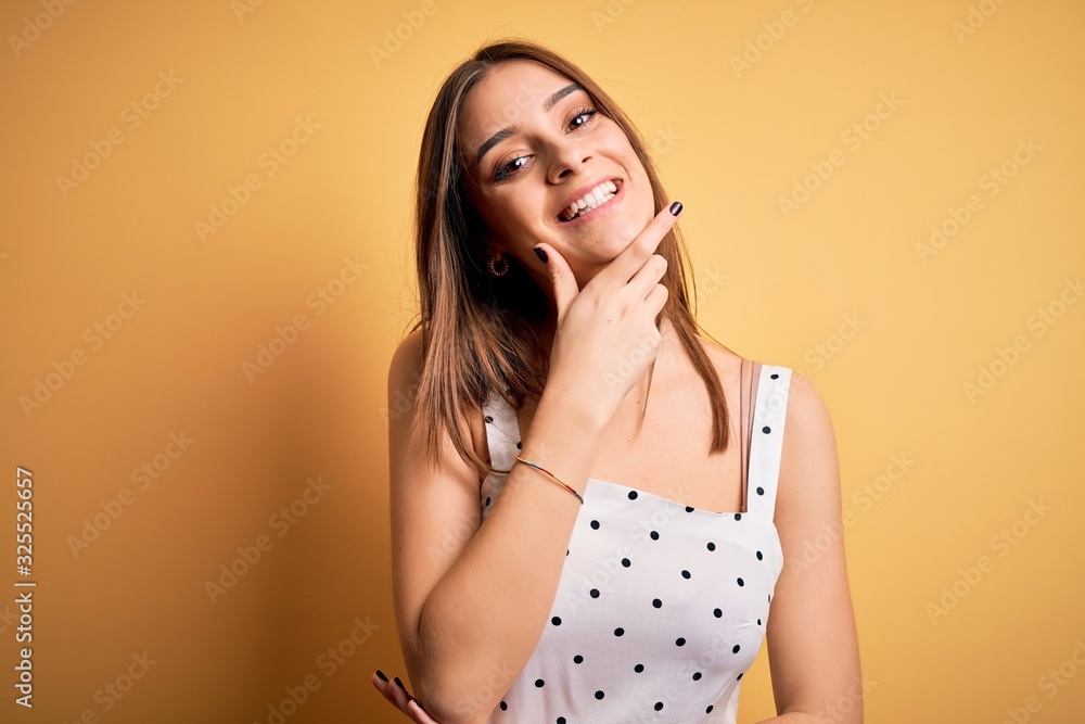 Fototapeta premium Young beautiful brunette woman wearing casual dress standing over yellow background looking confident at the camera smiling with crossed arms and hand raised on chin. Thinking positive.