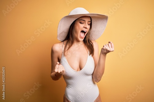 Young beautiful brunette woman on vacation wearing swimsuit and summer hat celebrating surprised and amazed for success with arms raised and eyes closed. Winner concept.