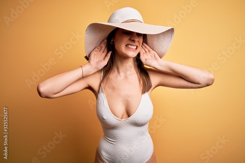 Young beautiful brunette woman on vacation wearing swimsuit and summer hat Trying to hear both hands on ear gesture, curious for gossip. Hearing problem, deaf
