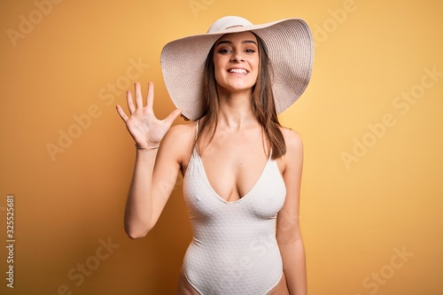 Young beautiful brunette woman on vacation wearing swimsuit and summer hat showing and pointing up with fingers number five while smiling confident and happy.