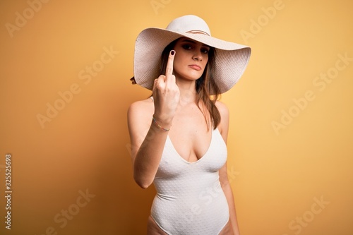Young beautiful brunette woman on vacation wearing swimsuit and summer hat Showing middle finger, impolite and rude fuck off expression